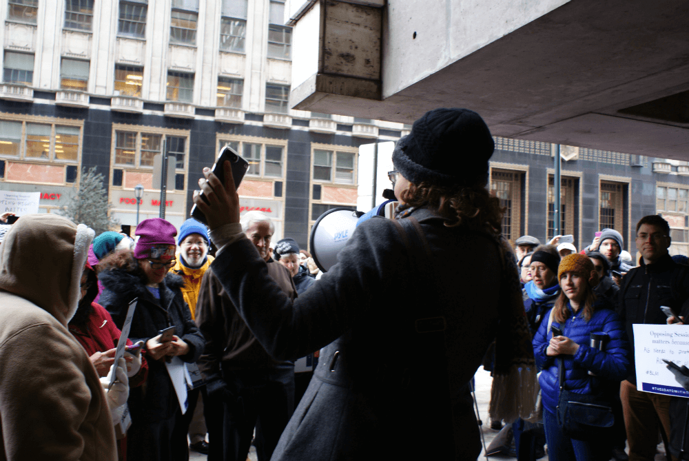 |<image-caption>
<p>Vashti Bandy holds her phone up to attendees at #TuesdaysWithToomey, and announces|Alexis Sachdev” title=”|<image-caption>
<p>Vashti Bandy holds her phone up to attendees at #TuesdaysWithToomey, and announces|Alexis Sachdev” /></div>
<p><!-- END scald=2783 --></div>
</div>
<p>“We want him to work for us, and we want him to listen to us,” Bandy said.
<p>Tom Paine Cronin, a lifetime Philadelphia resident and former leader of District Council 47, made his first appearance at Tuesdays with Toomey this week. He said Trump’s upcoming inauguration brought him out into the cold, and agreed with Bandy’s call for Toomey to lend an ear.</p>
<p>“Someone, a United States senator or any representative of the government, ought to be there to hear what people have to say, whether they agree with them or not,” Cronin said. “You’re supposed to be at least accessible to your constituents…The fact that [Toomey] just got re-elected, you would hope that you could influence him or open him up a little bit to be more receptive to people who have different opinions.”</p>
<p>Listening more was among the <a href=