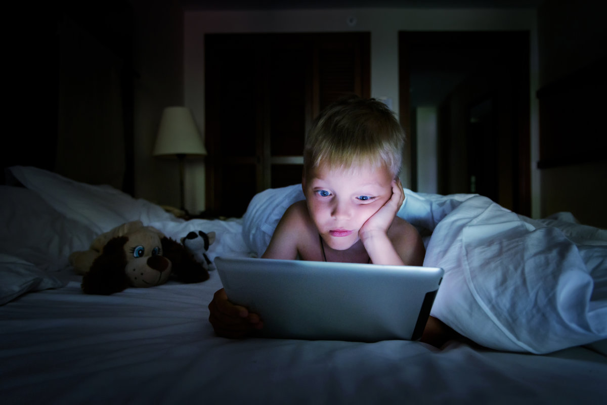 Satisfied kid boy using tablet to play playing learning application, lying under a white blanket in the bedroom at night, the blue light of the screen reflects his face.